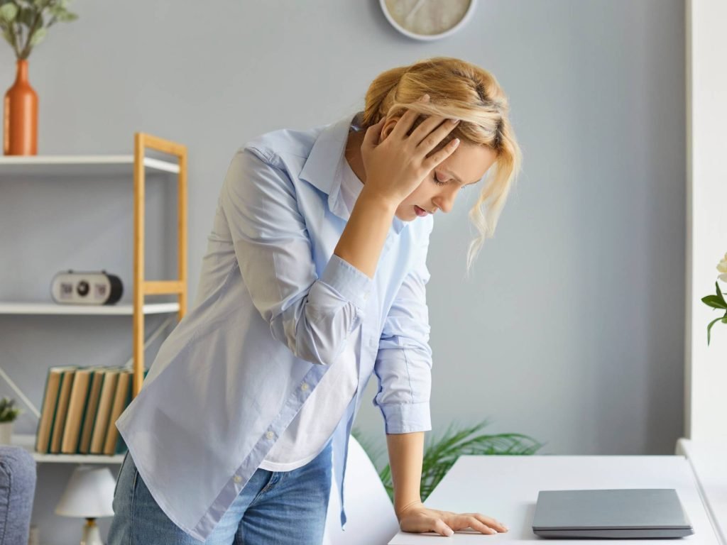 Women displaying signs of vertigo with hand on head in pain and holding onto desk for support.