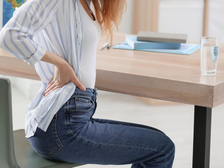 alt="A woman sitting on at her desk with lower back pain, a common symptom of sciatica and spinal misalignment."