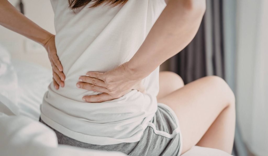 alt="A woman sitting on edge of bed with lower back pain, a common symptom of sciatica and spinal misalignment."